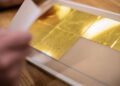 Close-up of a hand lifting a sheet of thin gold leaf squares placed on a beige backing paper.