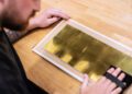 Person handling a large, rectangular sheet of gold leaf placed on cardboard, with both hands visible, at a wooden table.