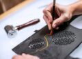 A person uses a tool to carve an intricate woodwork pattern with orange paint on a piece of black material with circular cutouts; a roller tool lies nearby.