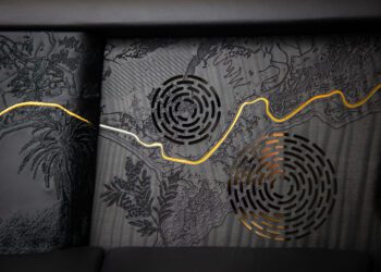 Black textured panel with circular speaker grilles and a wavy gold line, featuring embossed botanical patterns, detailed etching, and intricate woodwork inspired by Rolls-Royce craftsmanship.