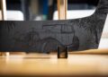 Close-up of a curved metal surface engraved with a Rolls-Royce vintage car and palm trees, standing upright on a wooden holder featuring intricate woodwork.