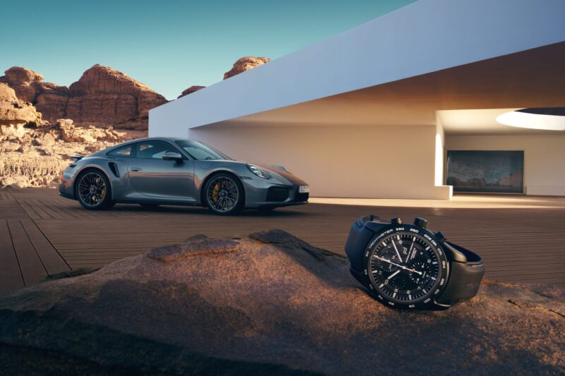 A silver Porsche Design sports car is parked outside a modern white house in a desert setting, with a Chronograph 911 Turbo S resting on a rock in the foreground.