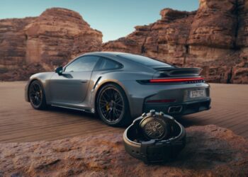 A luxury 911 Turbo S sports car is parked on a desert road with large rock formations in the background; a Porsche Design Custom-Built Chronograph wristwatch rests on a rock in the foreground.