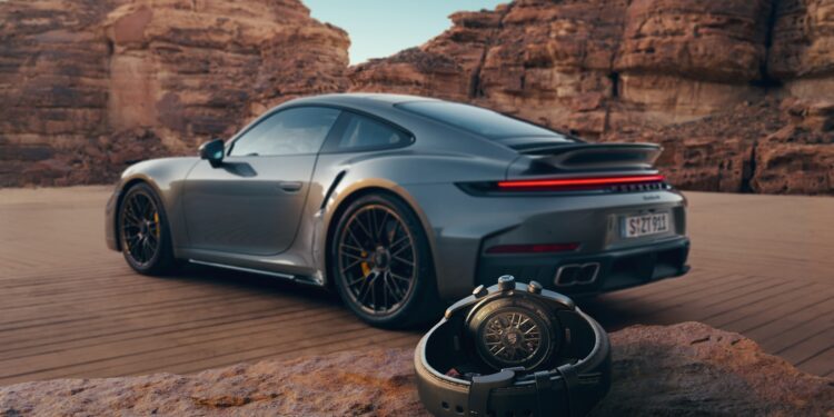 A luxury 911 Turbo S sports car is parked on a desert road with large rock formations in the background; a Porsche Design Custom-Built Chronograph wristwatch rests on a rock in the foreground.