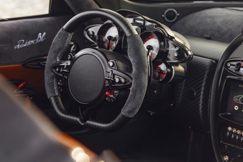 Close-up of a luxury sports car steering wheel with suede grip, analog gauges, and part of the infotainment screen—reminiscent of Pagani elegance set to shine as Pagani showcases four hypercar icons at the upcoming 9th Historic Minardi Day in Imola.