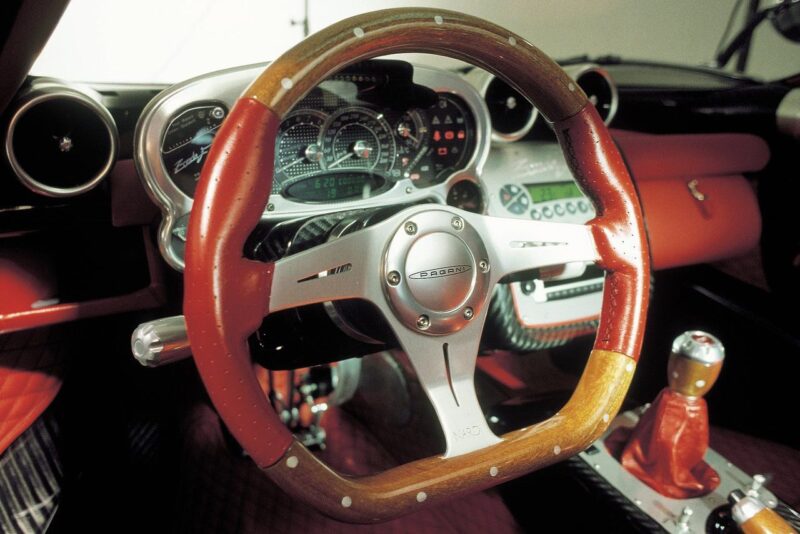 A close-up view of a sports car interior featuring a wooden steering wheel, analog gauges, and a gated manual shifter with a red knob—reminiscent of the iconic craftsmanship Pagani will showcase at the 9th Historic Minardi Day in Imola.