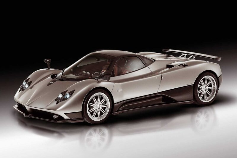 A silver sports car with aerodynamic design, large rear wing, and low profile, reminiscent of Pagani to showcase four hypercar icons at the upcoming 9th Historic Minardi Day in Imola, photographed on a reflective surface against a dark gradient background.