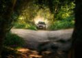 A black car drives on a dirt road in a forest, kicking up dust as it approaches a fork in the path surrounded by dense green foliage.