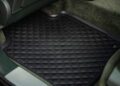 A black, quilted car floor mat with a rubber border is placed in the rear footwell of a vehicle with green carpeting.