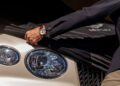 A person wearing a wristwatch pulls a black cover off the front of a Bentley car, revealing its headlights and grille.