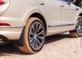 A white luxury SUV with distinctive alloy wheels is parked on sandy terrain, showcasing sand on the tires and ground—an ideal scene for featuring premium Bentayga accessories.