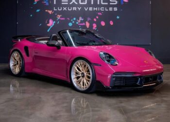 A pink Porsche 911 Turbo convertible sports car with gold alloy wheels is parked indoors under a sign that reads "1 Exotics Luxury Vehicles," showcasing one of the best models for sale.