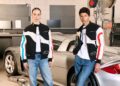 Two people in matching black, white, and colored racing jackets and blue jeans stand in front of a silver Porsche Carrera GT, part of an Exclusive Collection, in a garage setting.