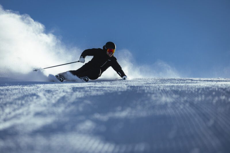A skier in black gear and red goggles, part of the Porsche ski collection, makes a sharp turn on a snowy slope, creating a spray of snow under a clear blue sky.