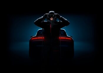 A person in dark clothing and a helmet stands in front of a Porsche sports car with illuminated rear lights, set against a dark background.