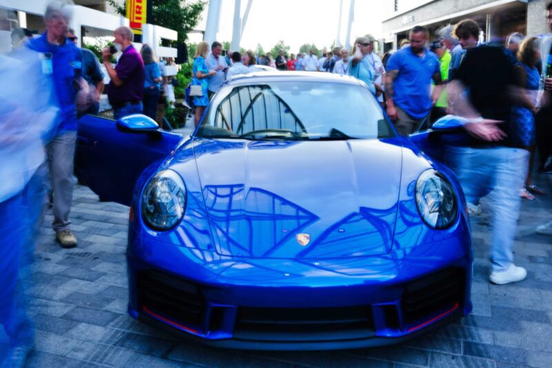 A blue Porsche sports car is parked outdoors, drawing the attention of a lively car community. Some members of the crowd appear blurred due to motion, capturing the excitement typical at Porsche Club of America gatherings.