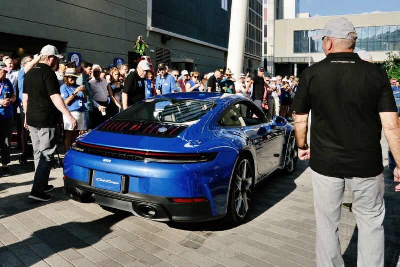 A blue Porsche sports car is parked on a city street, drawing a crowd of people taking photos and observing—a testament to the passion of the Porsche community.