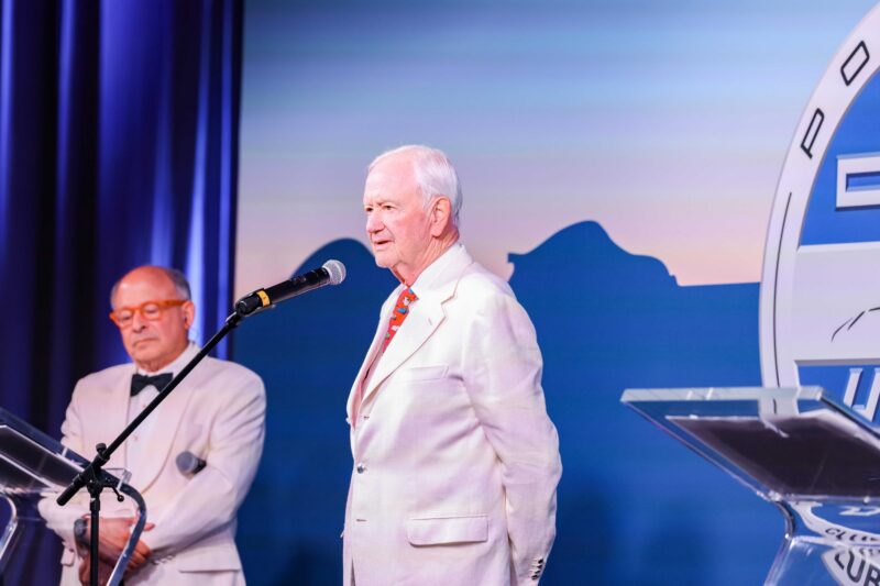 Two elderly men in white suits stand at podiums on a stage, celebrating a Porsche anniversary; one speaks into a microphone while the other stands in the background.