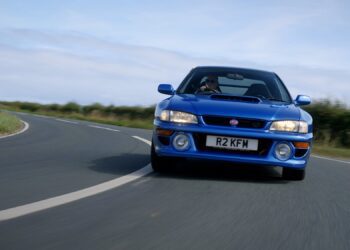 A blue Subaru 22B STi with the license plate "R2 KFW" is driving on a curved, paved road with greenery and a cloudy sky in the background, perfect for a Petrolicious Film Friday feature.