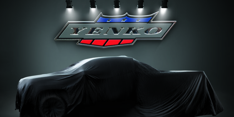 A car covered with a black cloth is displayed under spotlights, with a large Yenko logo mounted on the wall behind it.