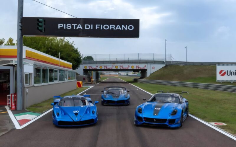 Three blue hypercars are parked side by side on a racetrack at the Fiorano Circuit, with the track's sign and structures visible in the background.