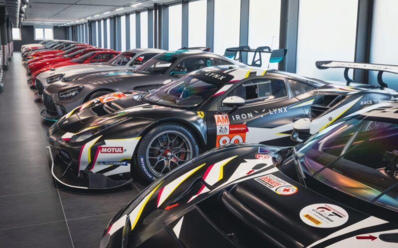 A row of high-performance sports cars, including hypercars with sponsor decals, are parked indoors in a modern, well-lit garage—evoking the prestige of an RM Sotheby’s Zurich auction.