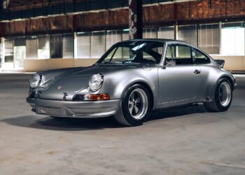 A classic silver Custom-Built Porsche 911 coupe is parked indoors on a concrete floor, with large windows and exposed beams in the background—a stunning example from The RSR Project.