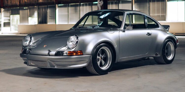 A classic silver Custom-Built Porsche 911 coupe is parked indoors on a concrete floor, with large windows and exposed beams in the background—a stunning example from The RSR Project.