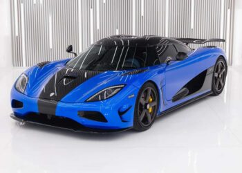A blue and black Koenigsegg Agera S with a sleek design is parked indoors on a white floor, set against vertical metallic wall panels—an eye-catching supercar that could be yours, now for sale.