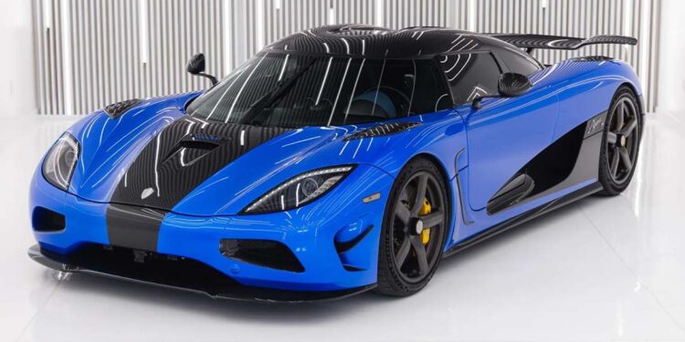 A blue and black Koenigsegg Agera S with a sleek design is parked indoors on a white floor, set against vertical metallic wall panels—an eye-catching supercar that could be yours, now for sale.
