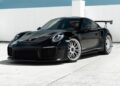 A black Porsche GT2 RS with chrome wheels is parked on a concrete surface against a white wall under a blue sky.