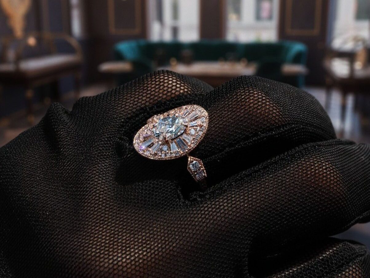 A hand wearing a black glove holds a marquise-cut diamond ring with halo and side stones—an AviNika Jewelry masterpiece—gleaming under chandelier light, destined to become one of your timeless heirlooms.