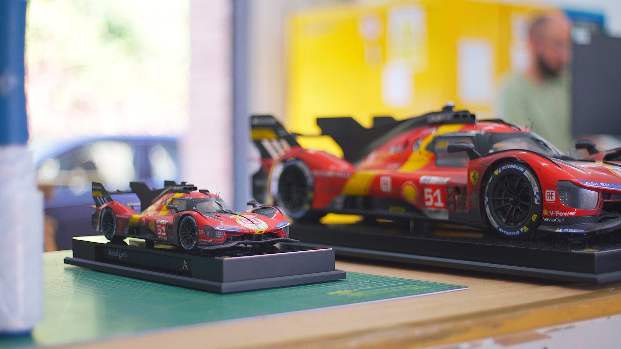 Two detailed Amalgam Collection scale models of red and yellow race cars are displayed on a table, with a blurred background showing a person and a yellow object—a true Petrolicious miniature scene.