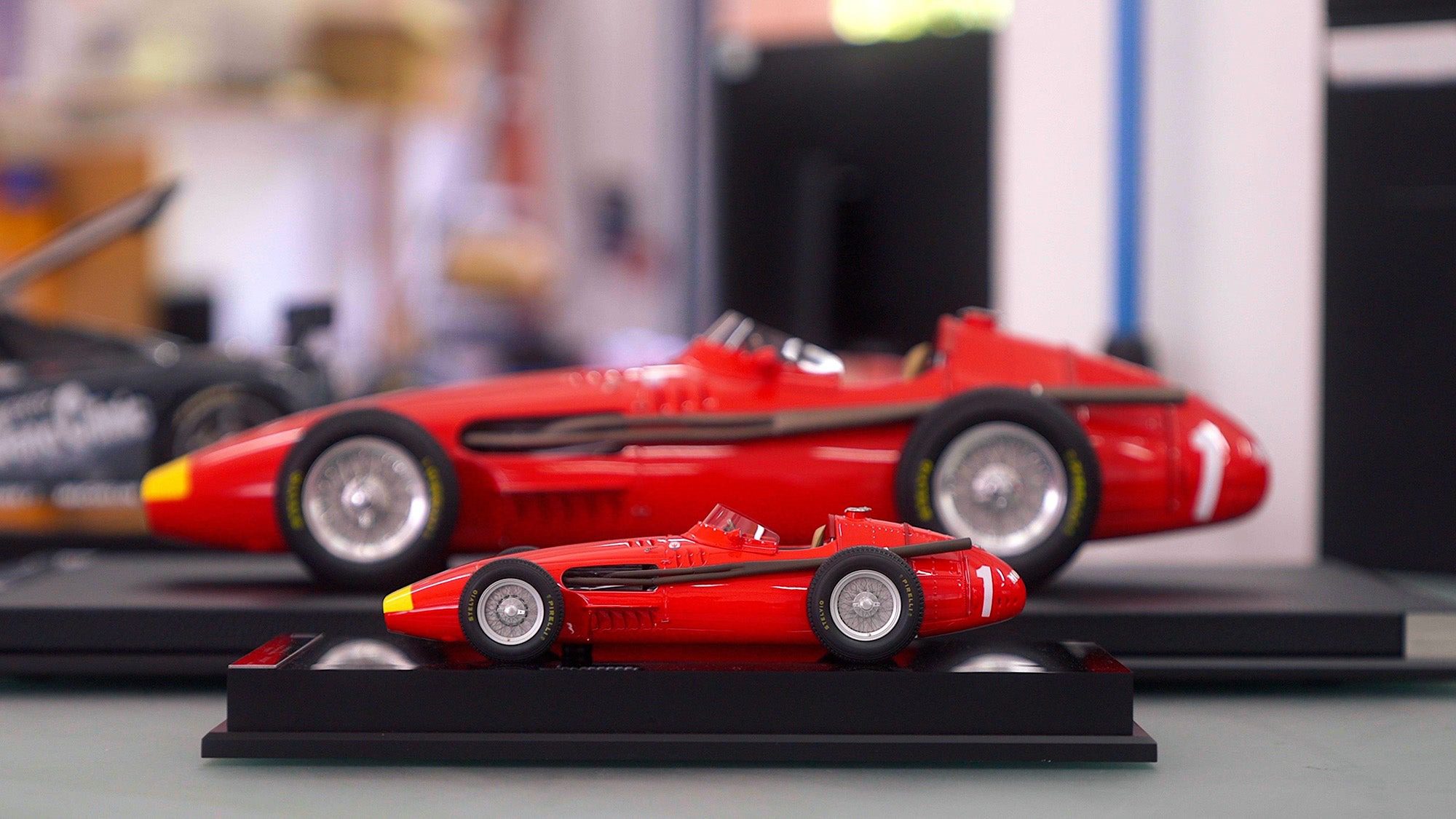 A close-up of two red vintage race car models, one small and one large, displayed on black stands indoors—a striking miniature legacy echoing the craftsmanship of the Amalgam Collection.