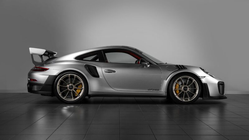 A silver 991 Porsche 911 GT2 RS sports car is parked indoors on a tiled floor, viewed from the side against a plain gray wall—a stunning example of value in a model known for its skyrocketing desirability.