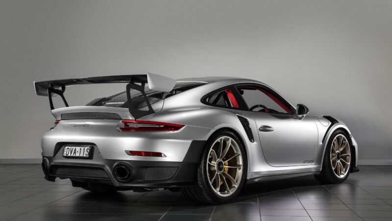 A silver 991 Porsche 911 GT2 RS sports car is parked indoors on a tiled floor, viewed from the rear side, showcasing its value with a large rear wing and striking gold wheels.