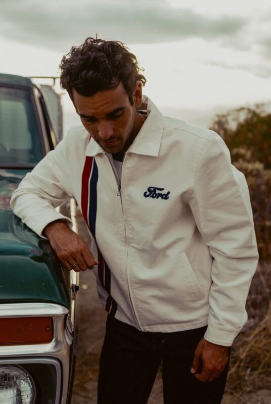 A man wearing a white Ford x Brixton jacket with red and blue stripes stands next to a green vintage vehicle outdoors.