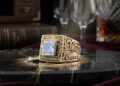 A gold ring with an ornate design and a large square gemstone is displayed on a marble surface, embodying the rare legacies of AviNika Jewelry, with wine glasses, a book, and a decanter in the background.