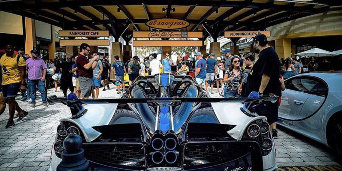 A crowd gathers around luxury sports cars, including a Pagani Huayra, outside The Colonnade shopping area on a sunny day.