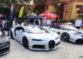 Several high-end sports cars, including a white Bugatti, are parked in front of vendor tents at an outdoor car show with people walking around.