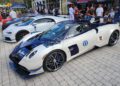 A blue and white Pagani Huayra with racing decals is parked among other luxury sports cars at an outdoor car show with people observing.