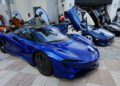 A row of blue luxury sports cars with scissor doors open are displayed outdoors at a car show, with several people walking nearby.