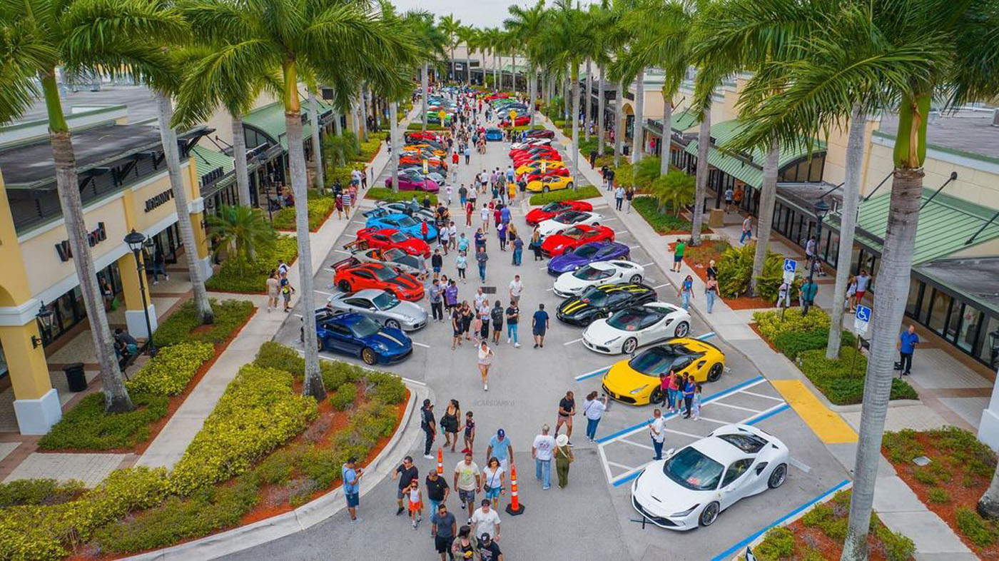 Aerial view of a car show with crowds walking among rows of colorful sports cars parked along a palm tree-lined street between shopping centers.