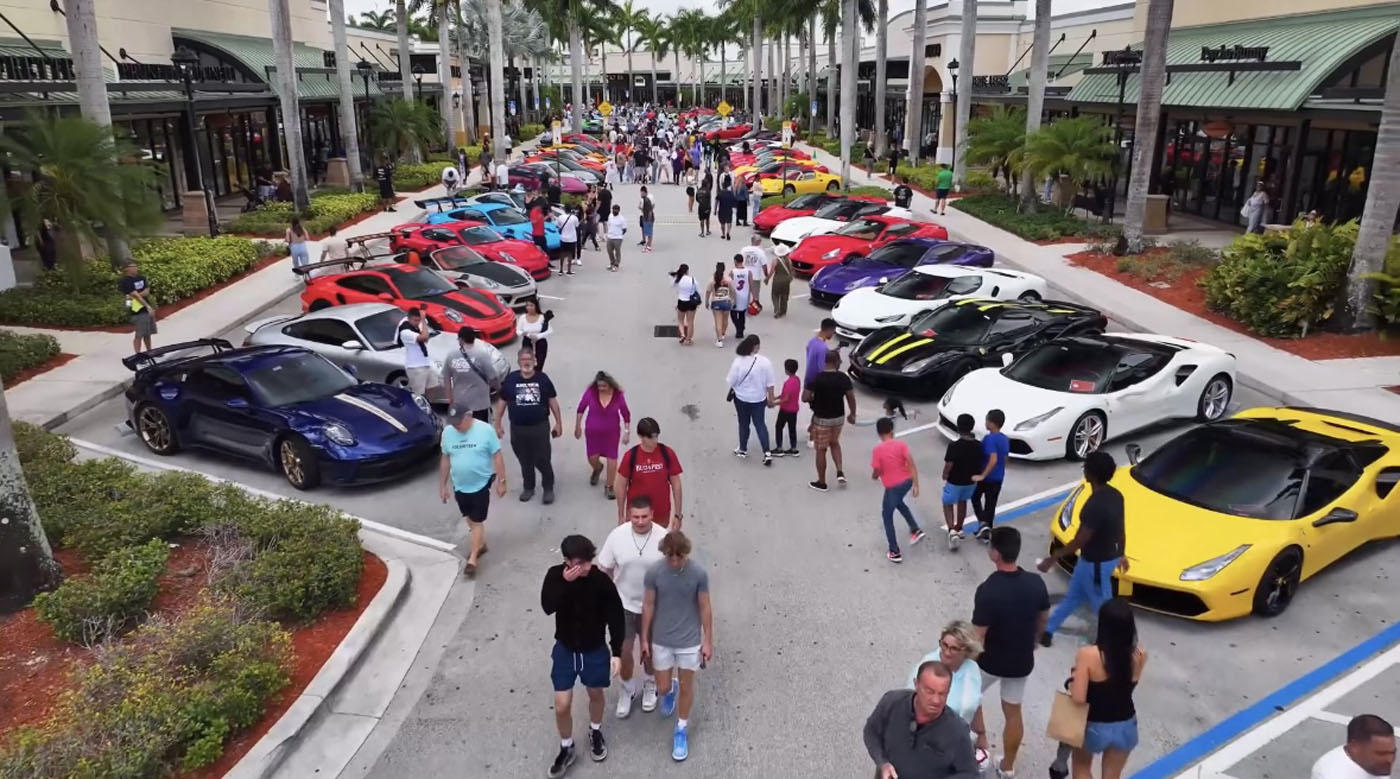 A crowd walks among rows of colorful sports cars parked outside shops at an outdoor car show, with palm trees lining the street.