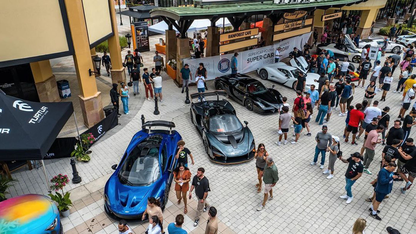 A crowd gathers around several luxury sports cars displayed outdoors in a plaza with shops and vendor booths.