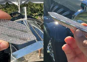 A hand holds a diamond-encrusted car hood ornament shaped like stylized wings, inspired by origami engineering, with a classic car in the background.