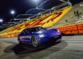 A blue Porsche Taycan Turbo GT drives on a racetrack at night, with empty yellow and red stadium seats in the background, reminiscent of scenes from Sydney Dragway where quarter-mile records are set.