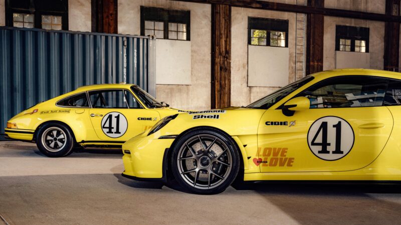Two yellow Porsche 911 S/T race cars with number 41 decals are parked indoors. The cars, adorned with sponsor logos and black racing stripes, embody the spirit of a true racing legend.