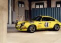 A yellow vintage Porsche 911 S/T race car, number 41, with Shell and Michelin logos, stands as a legend parked inside an industrial warehouse near a blue shipping container.
