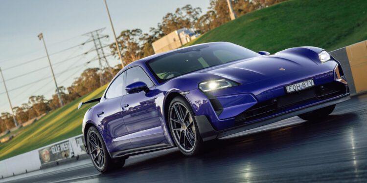 A blue Porsche Taycan is parked on a racetrack with barriers and a grassy hill in the background.